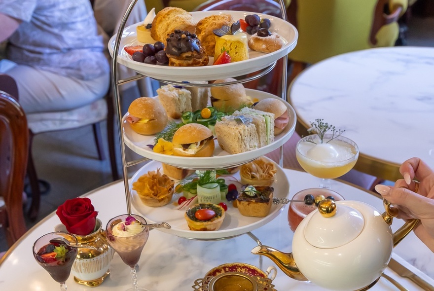 A three-tiered high-tea offering with cakes and small savoury bites, someone pouring tea from a pot. 