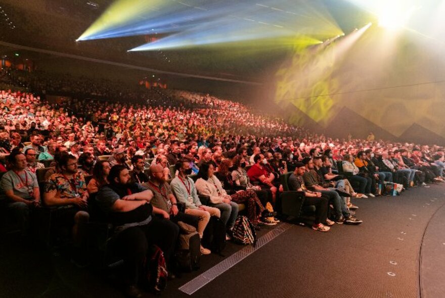 Seated audience in a large auditorium, with ranked rows of seating and laser lights overhead.