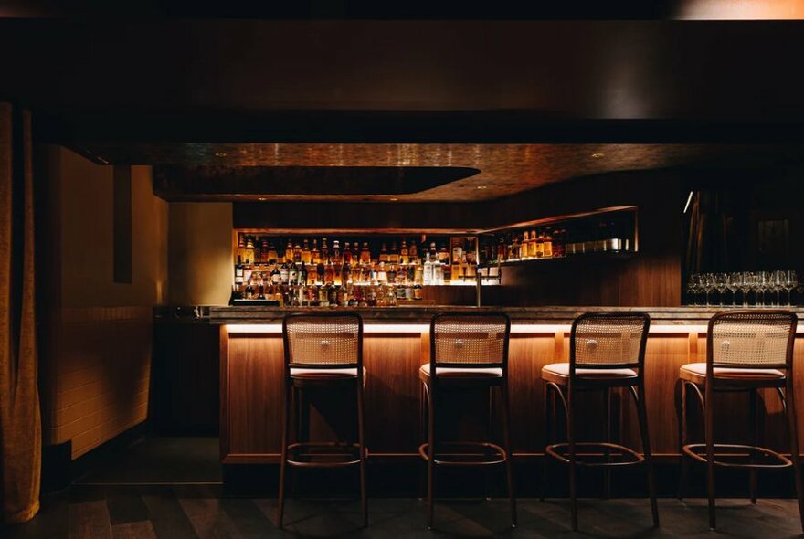Dimly lit basement bar at Central Club Hotel with four high stools perched at a bar, many bottles of spirits on the shelves behind.