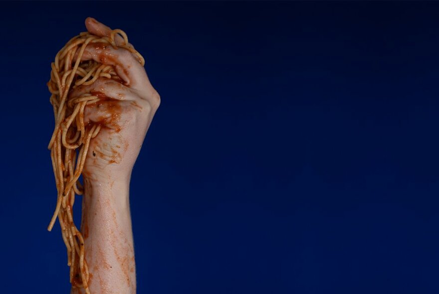 A raised hand holding tomato sauce-covered spaghetti in front of a dark blue background.