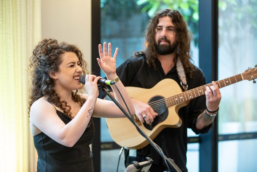 Two people, a singer and a guitarist, performing in an indoor space near a large window.