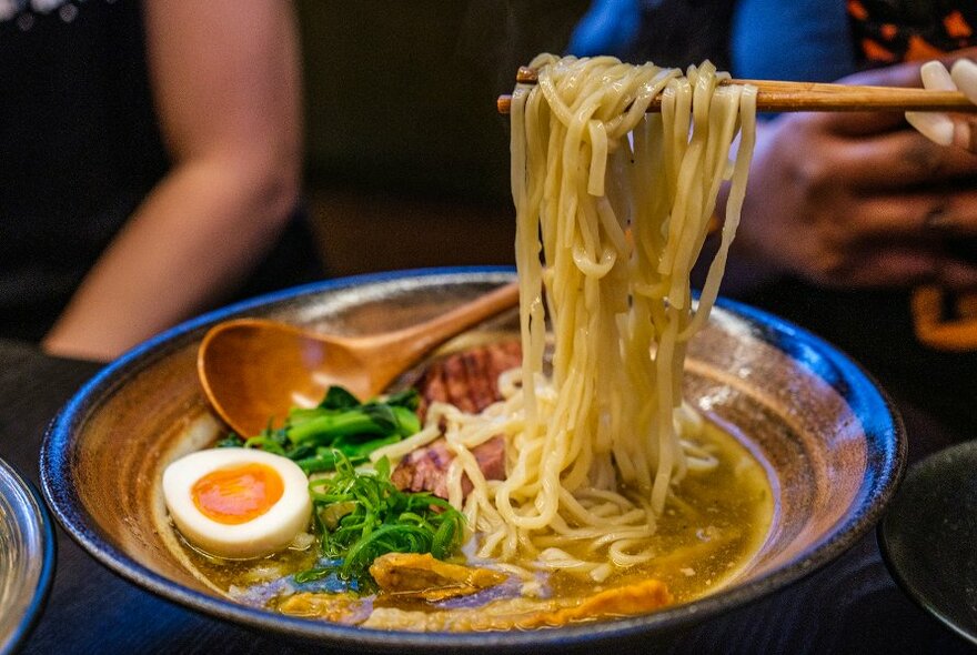 Brown bowl of ramen with chopsticks pulling out noodles. The bowl is topped with pork, greens and half an egg.