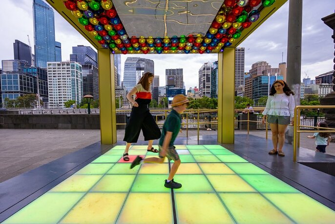 A child and woman play on a light-up dancefloor in a structure with a multi-coloured ceiling, with city buildings in the background.