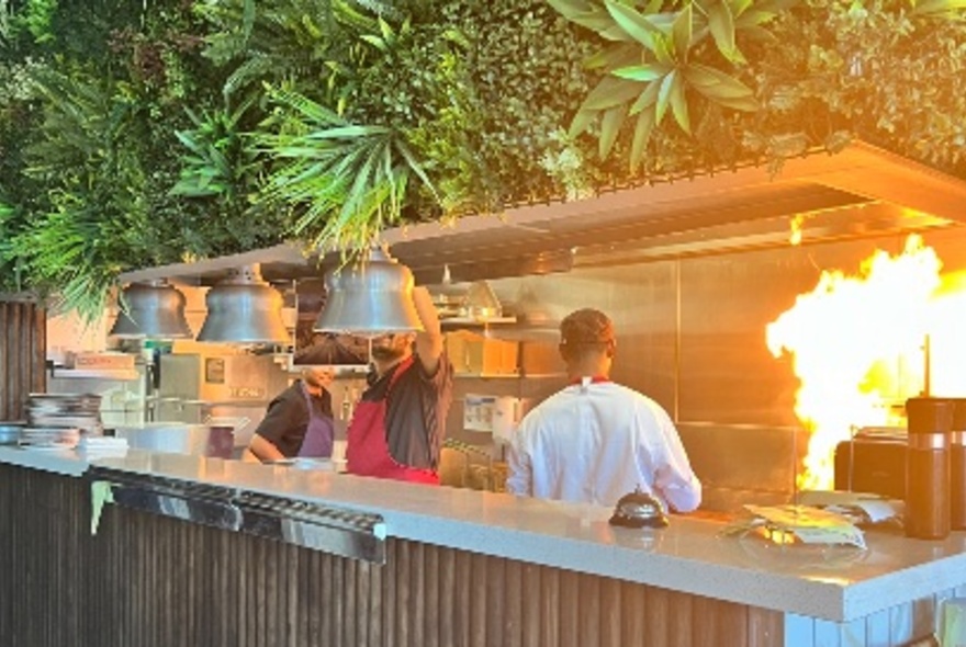 Flames bursting from grill as staff work the pass in a restaurant with overhead wall of plants.
