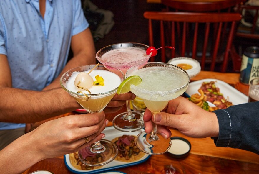 Three people doing cheers with three Mexican cocktails above a wooden table laden with delicious-looking Mexican food plates.