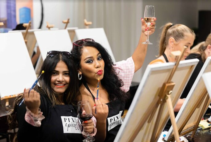 Two people sipping wine seated in front of an easel.
