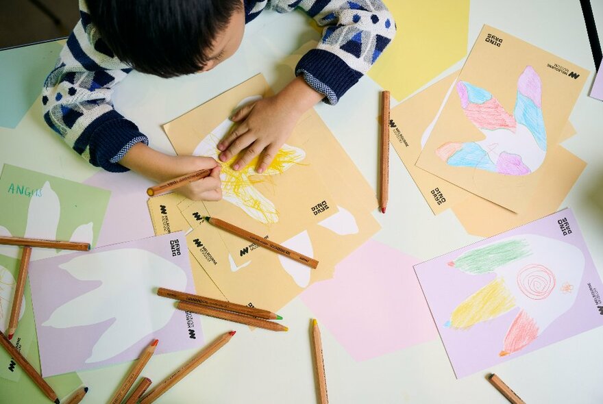 Young child colouring in with pencils on paper cutouts of dinosaur feet.