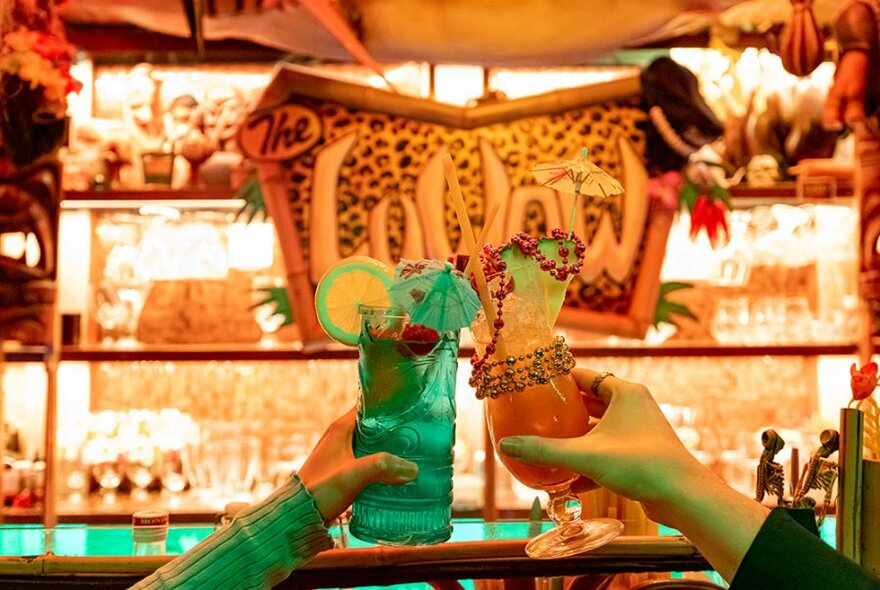 Two people holding up colourful cocktails with fruit, umbrellas and beads around them.