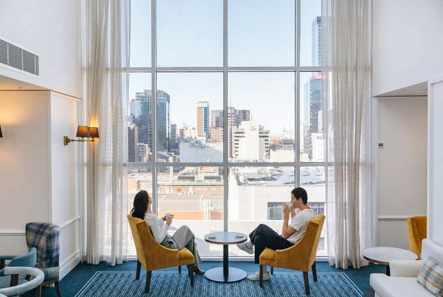 A couple seated in club armchairs inside a spacious hotel club lounge, with a view of the city skyline through large floor to ceiling windows. 