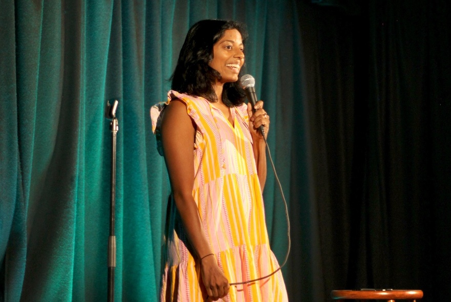 Performer standing on a small stage, talking into a microphone she holds in her hand.