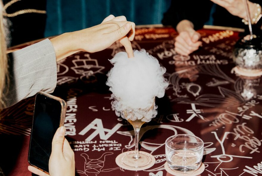 A person putting their finger in a cocktail with a fairy-floss / cloud-like top on a table with a graffiti pattered red tablecloth. 