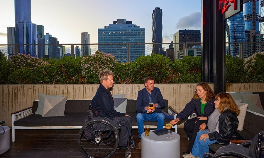 A group of friends drinking beer and cocktails on a rooftop overlooking the city skyline. Two of the group are sitting in wheelchairs.