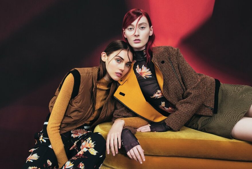 Two models in autumnal shades lounging on plush, mustard-coloured lounge.