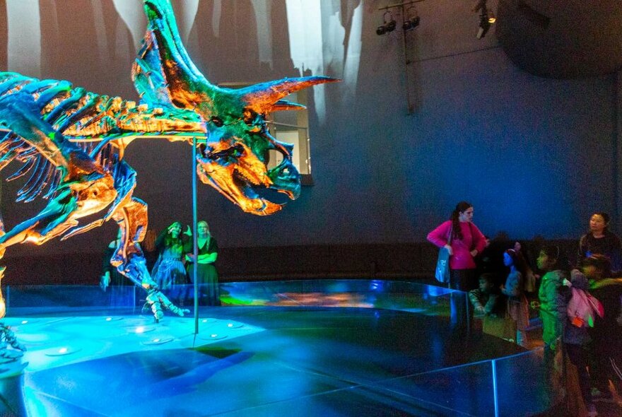 An illuminated dinosaur model under spotlights, with adults and children looking on.
