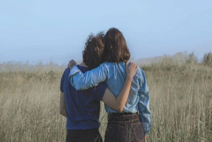 The back of two people, with one arm around each other, standing in a dry field. 