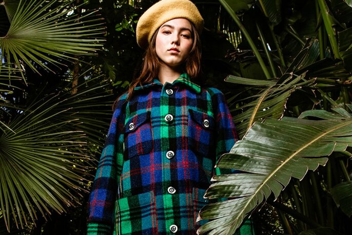 Model wearing a blue, green and black coloured and patterned coat and a pale orange beret on her head, standing outdoors among large palm plants.