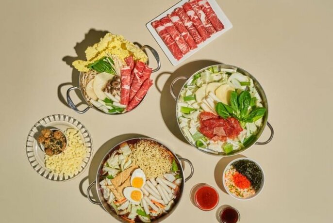 Looking down at a table of three large pans filled with fresh slices of meat, vegetables, boiled eggs, and noodles, with smaller plates of food around it.