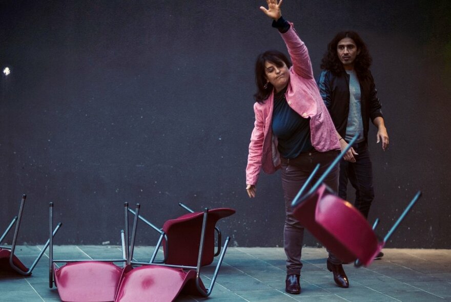 An actor wearing a pink jacket, hurling red chairs on a stage while an onlooker observes.