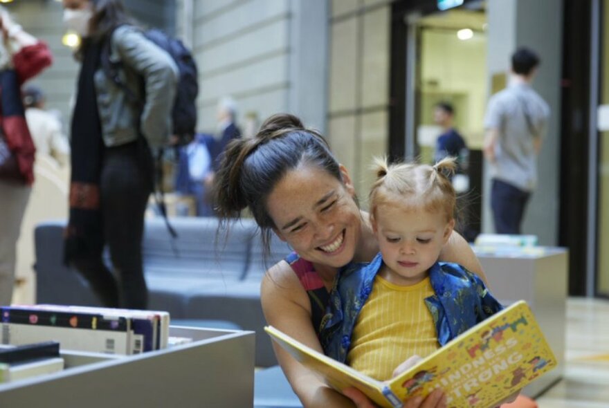 A toddler sitting on their mother's lap, both of them smiling and looking at a picture book, in the open space of a library.