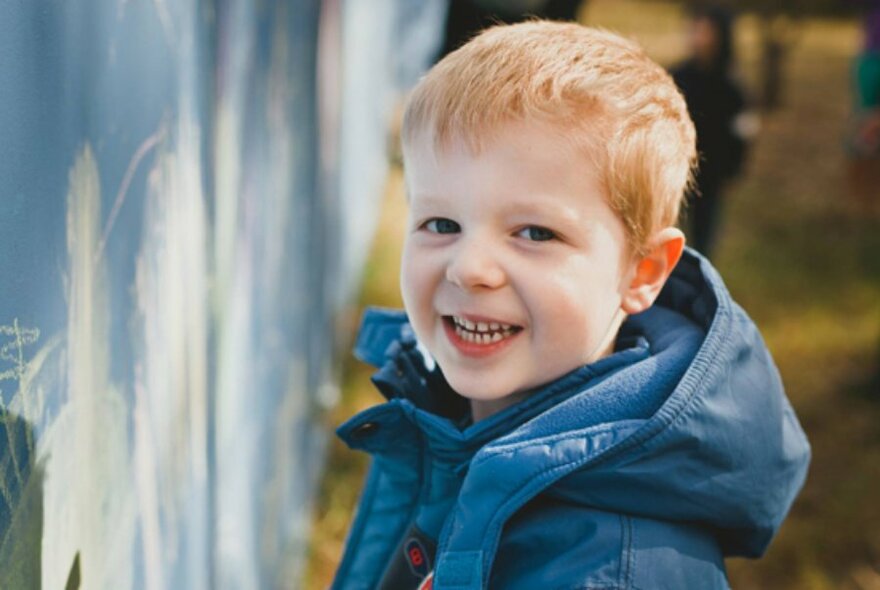A young red-haired boy in a blue coat smiling beside a painted wall.
