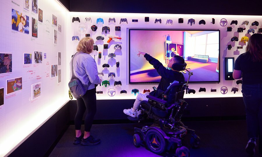 A person using a wheelchair pointing at a wall of video game controllers in a gallery.