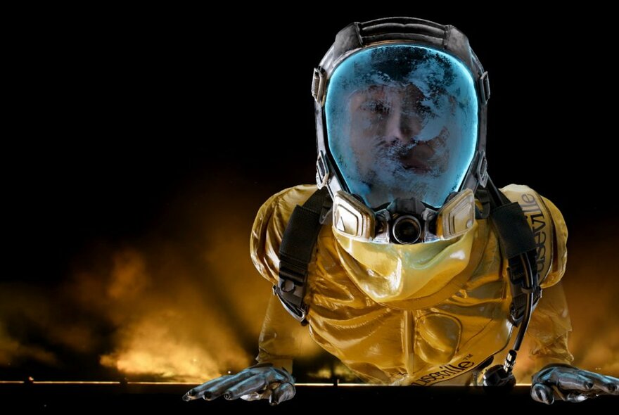 Person wearing a yellow spacesuit and space helmet, their face barely decipherable under the interior fog of the glass front of their helmet, their gloved hands resting on a panel in front of them, backlit with a yellow light.