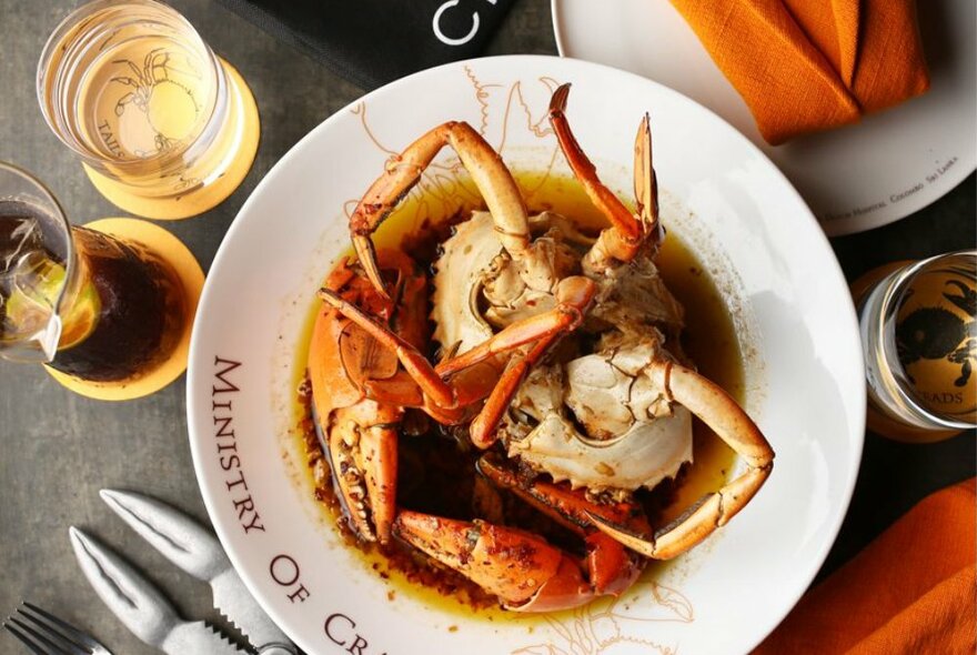Plate of whole mud crab in a sauce, on a table with glasses of wine, napkin and a crab-cutting tool.