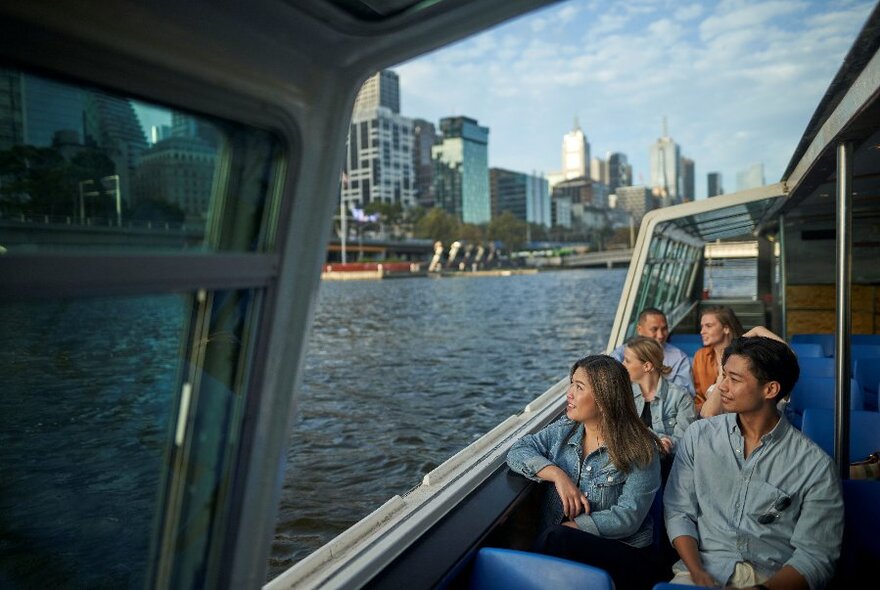 People seated in a river boat with outside seating, passing city buildings.
