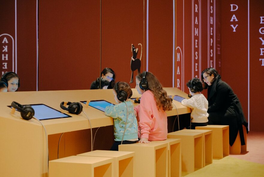 Children wearing headphones and interacting with screens that are set into a wooden desk console inside a large exhibition space.