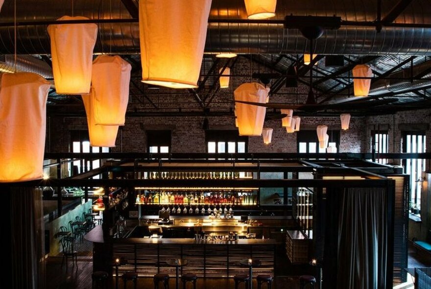 Glowing fabric lanterns hanging from the beams of a wooden roof above a backlit bar.