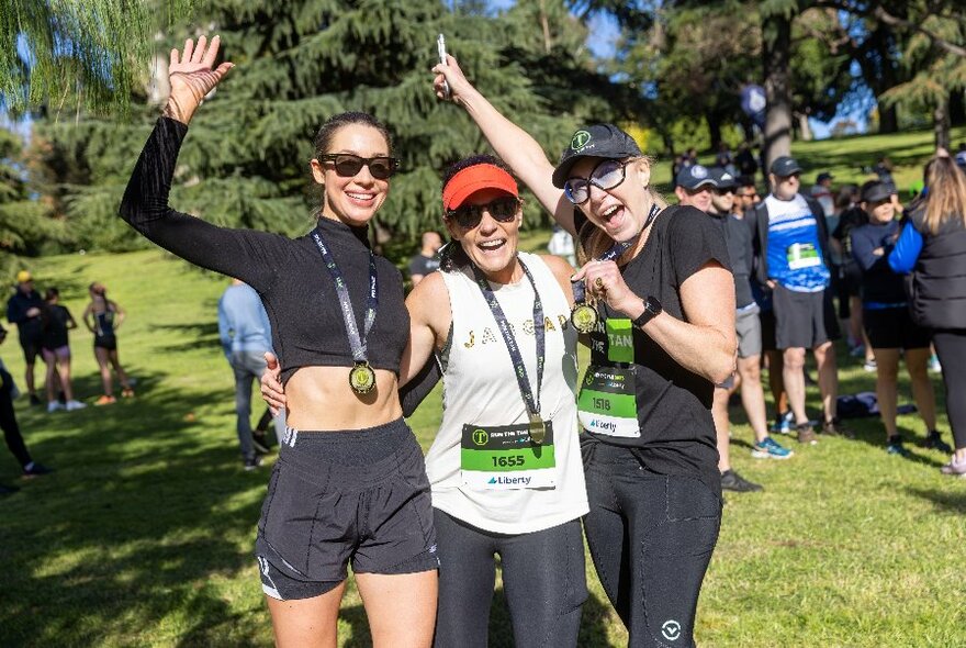 Three people hugging and cheering each other one at the end of a fun run, standing in a park, wearing their completion medals. 