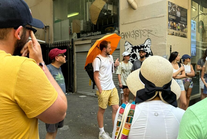 A guide holding an orange umbrella standing on a laneway corner and leading a small group of people on a walking tour of the city.
