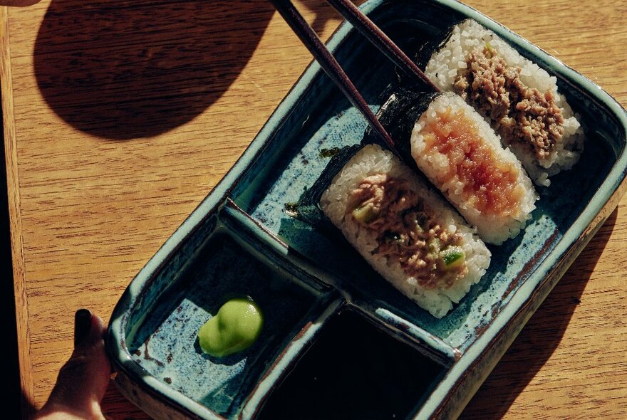 Three pieces of onigiri in a blue earthenware bento box with wasabi, and chopsticks.