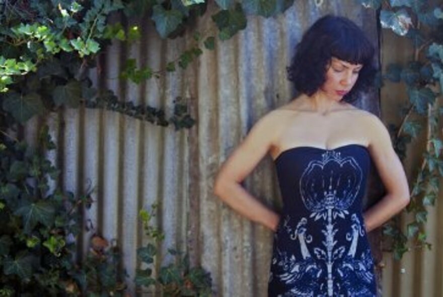 Woman wearing a strapless blue dress, looking down, while standing against a corrugated-iron wall.