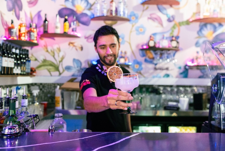 A bartender proudly holding forth a margarita while standing behind a bar with floral patterns on the wall.