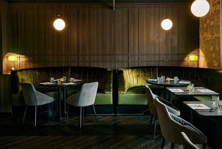 Inviting interior of a restaurant, with green velvet banquette seats, small tables, grey velvet club chairs, soft wall furnishings and hanging spherical lights.