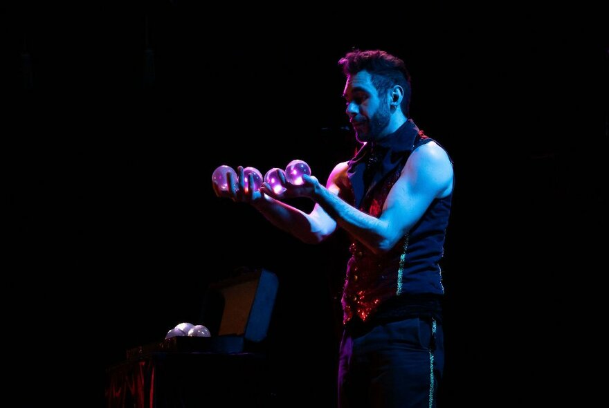 Juggler performing on a dark blue-lit stage holding four glass balls.