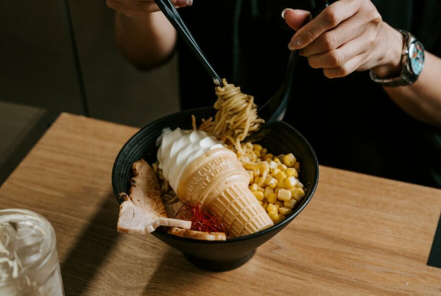 A bowl of ramen with corn and a soft serve ice cream in the middle, a person picking up the noodles with chopsticks. 
