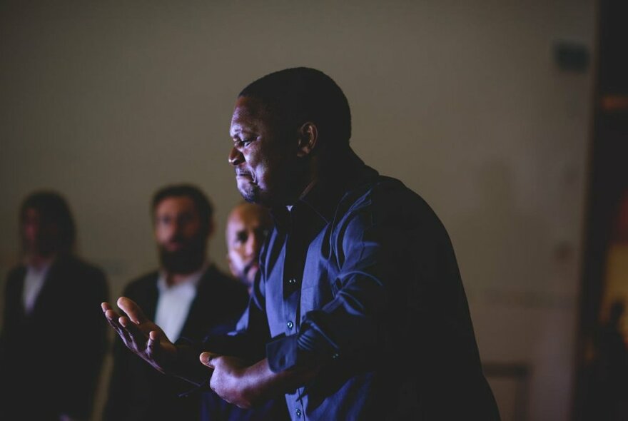 An Indigenous man standing and presenting in front of a group in a dimly-lit room. 