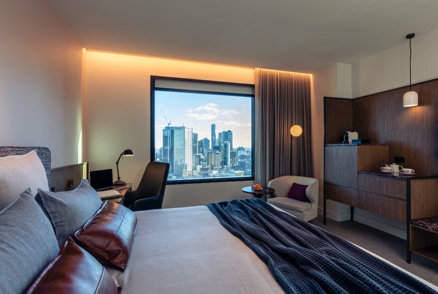 A hotel room at Next Melbourne with a large picture window showing the city skyline, a king size bed, and soft lights.