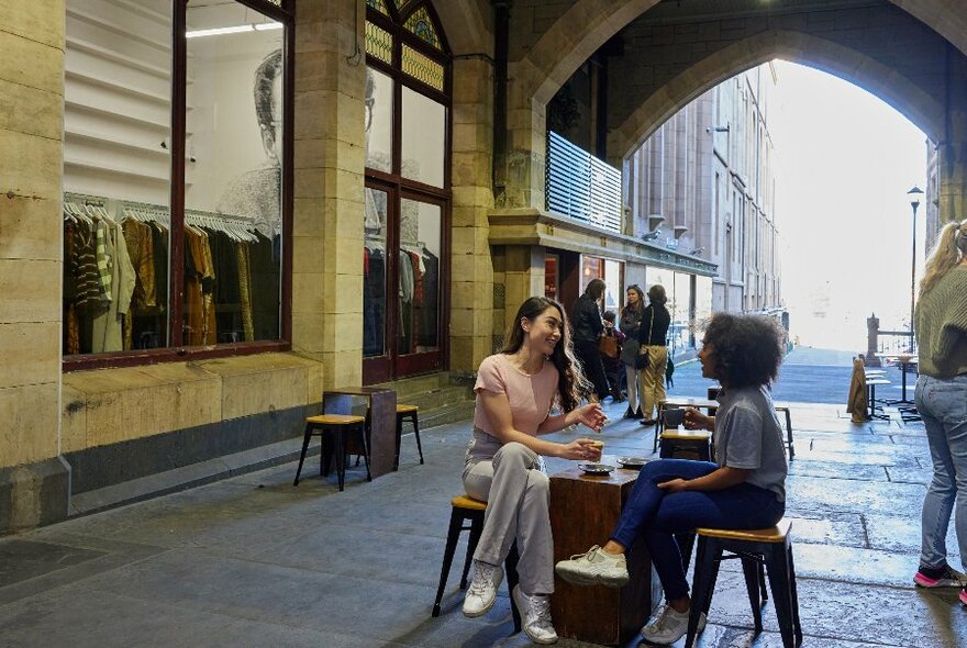 Two women are sitting at a table in a laneway with an arched ceiling drinking coffee.