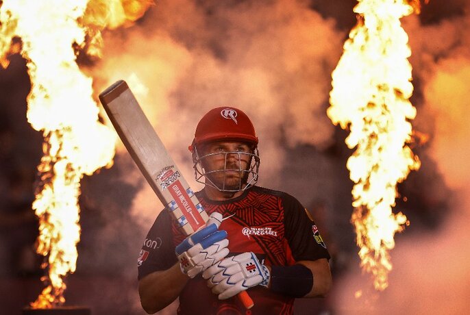 A cricketer holds his bat high surrounded by flames. 