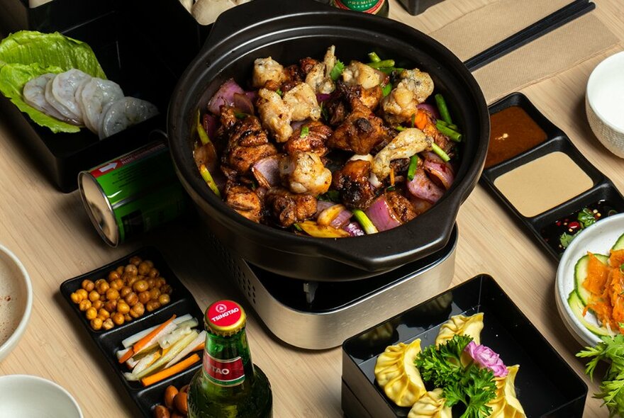Claypot of chicken and vegetables cooking on a burner on a dining table, with side plates of garnishes and condiments arranged around it.