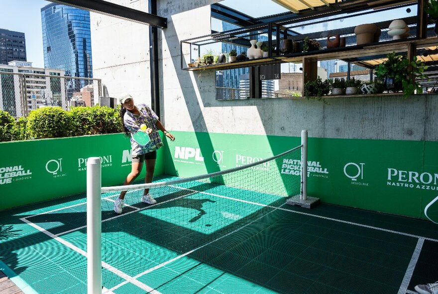 A player having a hit on the pickleball court at the rooftop bar at QT Hotel, Melbourne, with the city skyline in the distance.