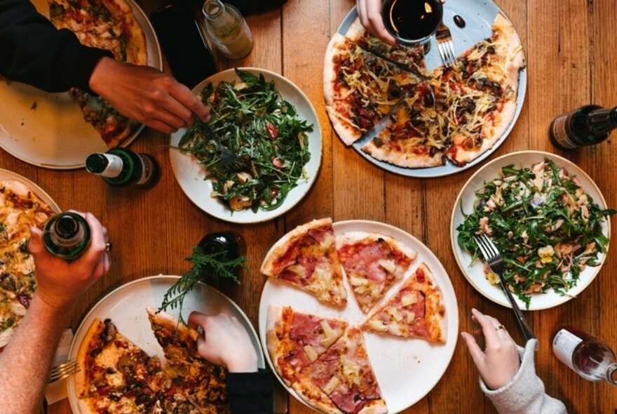 Pizzas and salads on a table, with hands reaching out for slices.