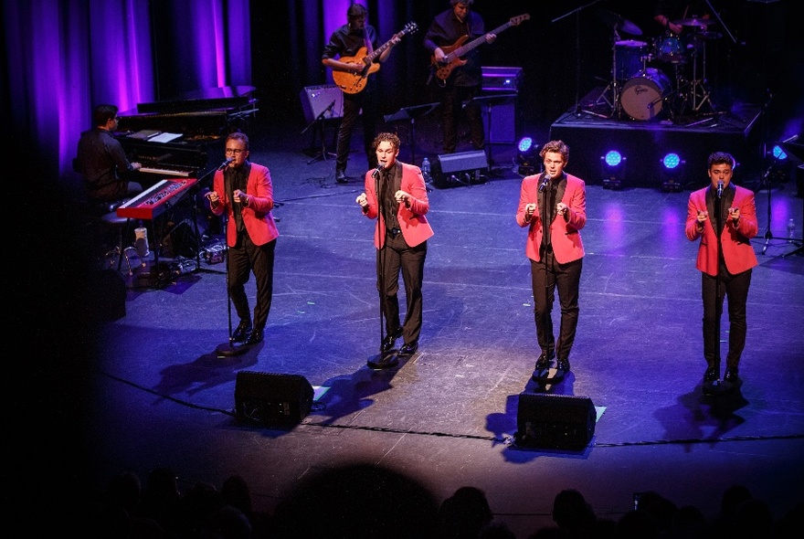 Four men in identical pink jackets and black shirts singing on stage and holding their hands in the same position. 