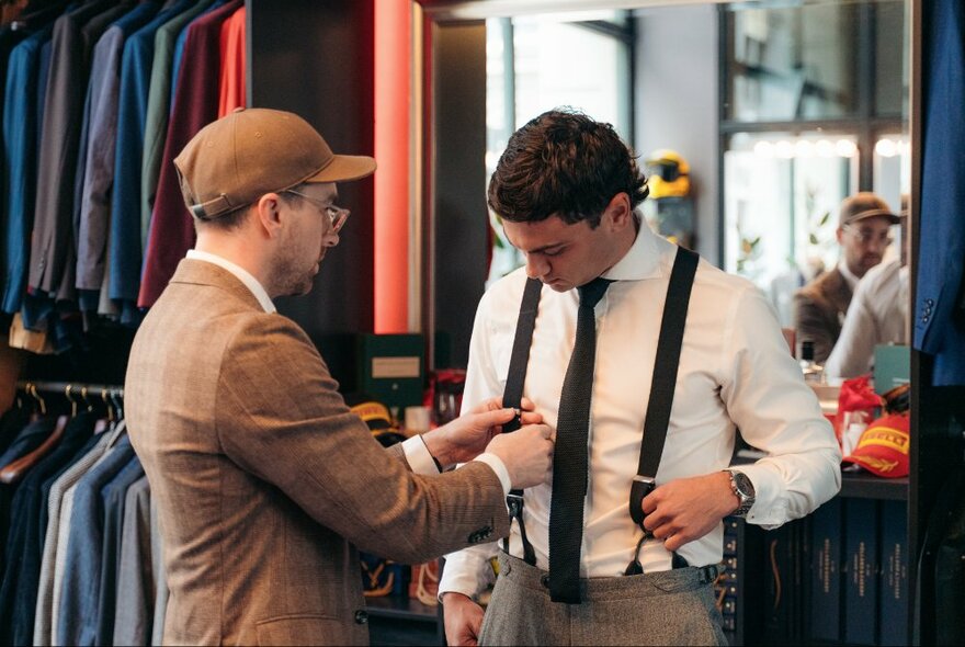 A tailor fitting braces onto a suit worn by a client in a men's fashion store.