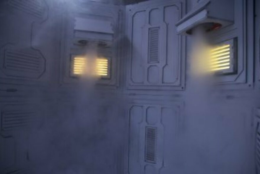 Inside a white-grey room with mist or smoke coming out of the wall vents.