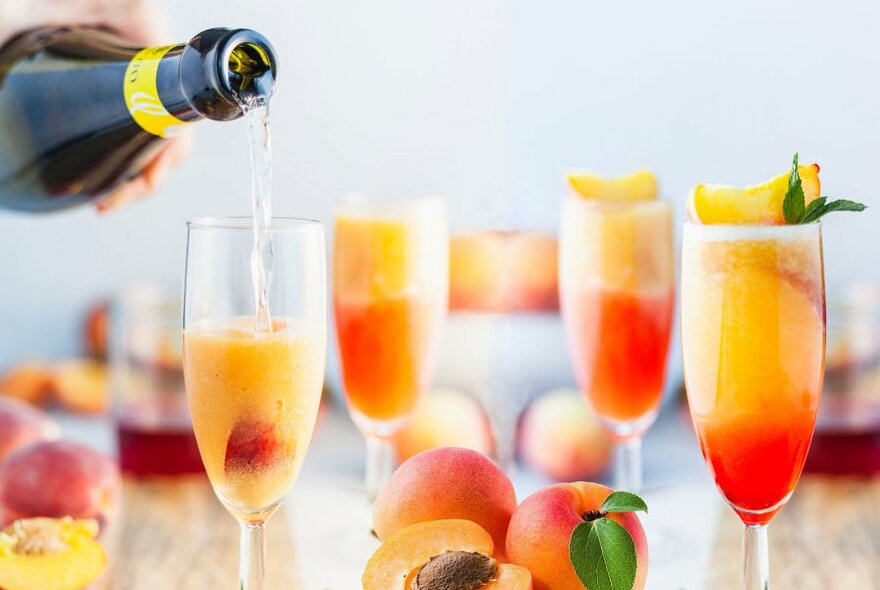 Champagne from a bottle being poured into flute glasses with fruit, other glasses and apricots in view on the table.