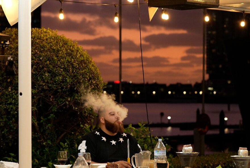 A man smoking a hookah at a restaurant table with a sunset view of Docklands in the background.